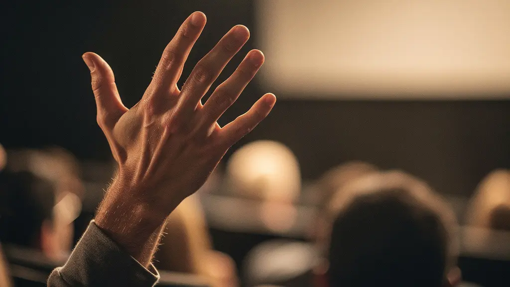 Close-up of a raised hand in a dimly lit cinema auditorium during a film festival panel discussion, with warm stage lighting illuminating the gesture against a blurred audience background.